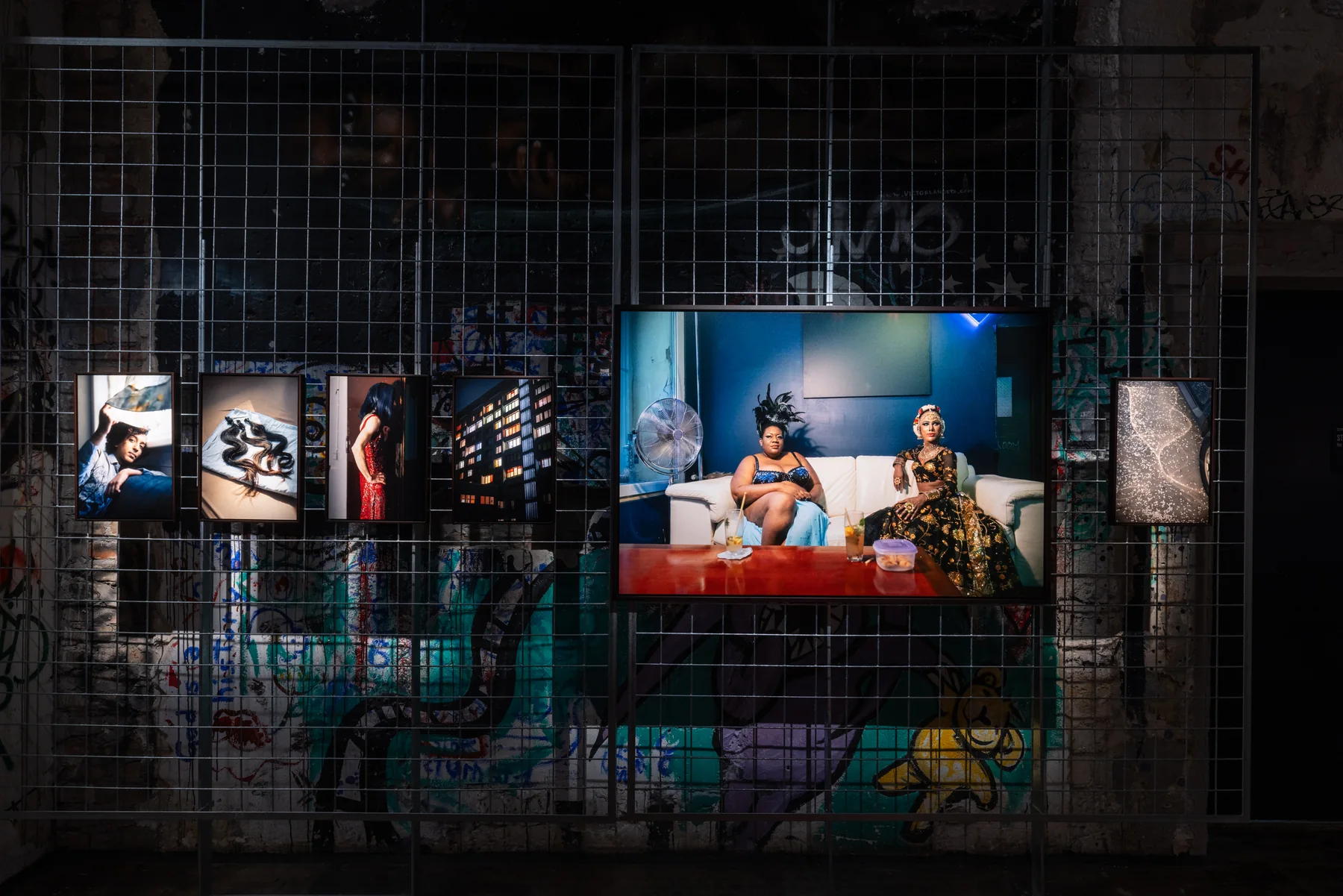 Installation view: a wall of prints centred on two figures seated on a sofa.
