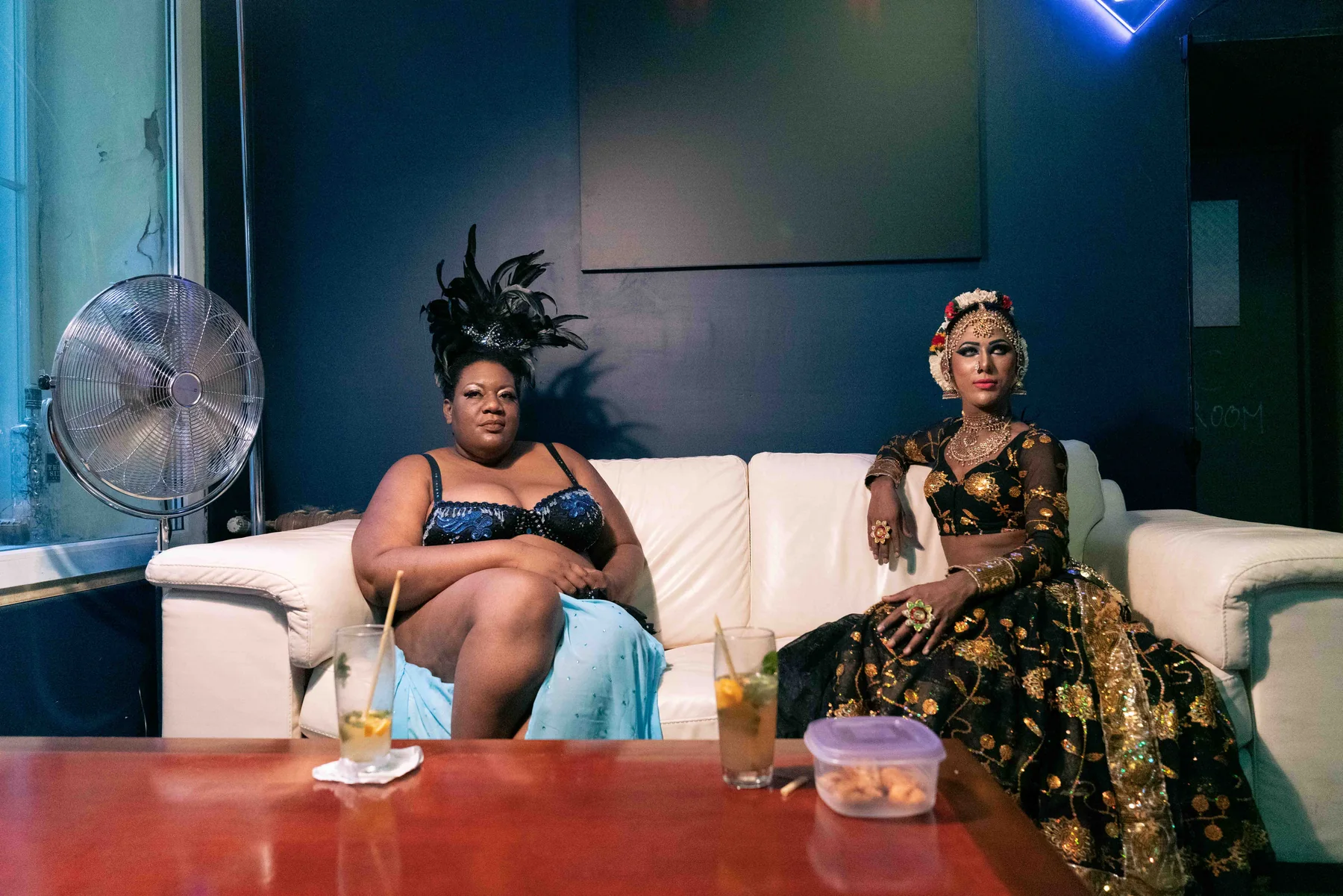 Two people in evening gowns sit on a white sofa, drinks on the table before them.