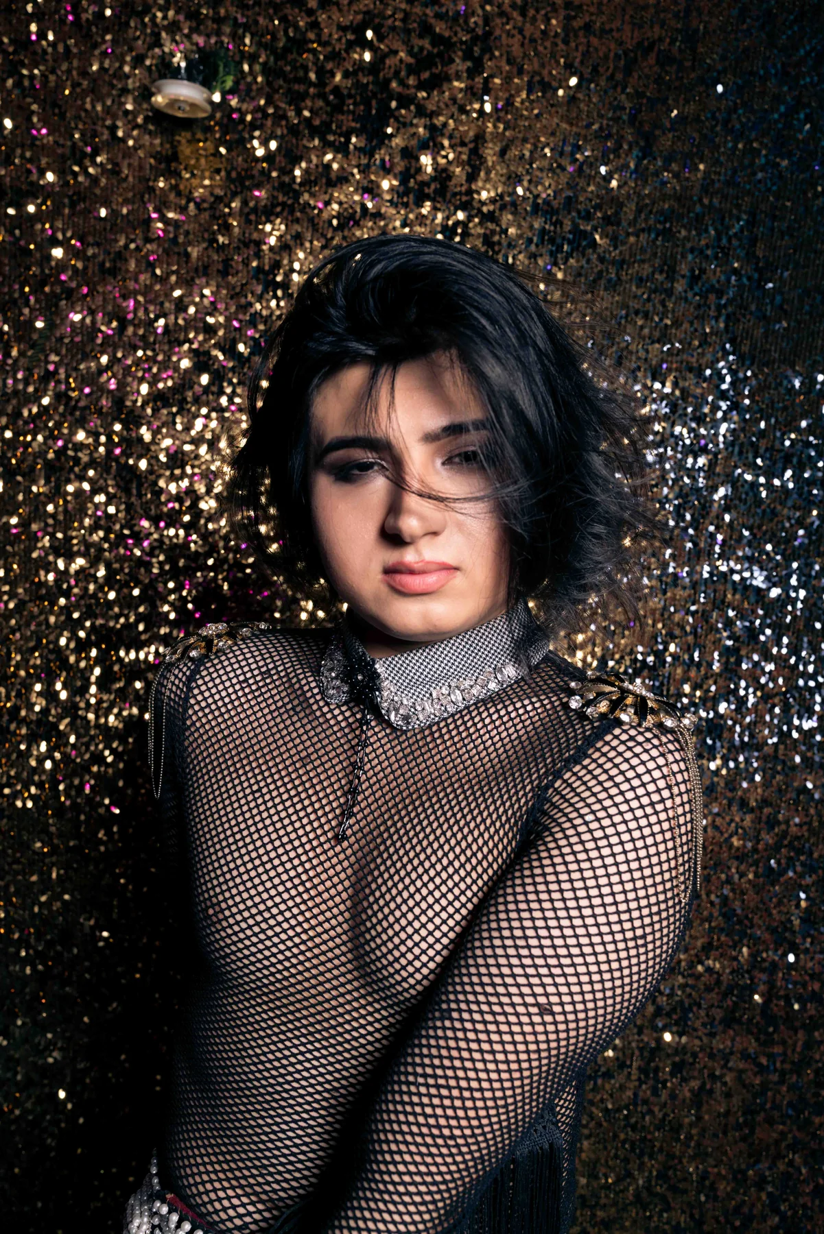 A person in a black mesh top stands against a dark wall flecked with light, arms folded across their chest.