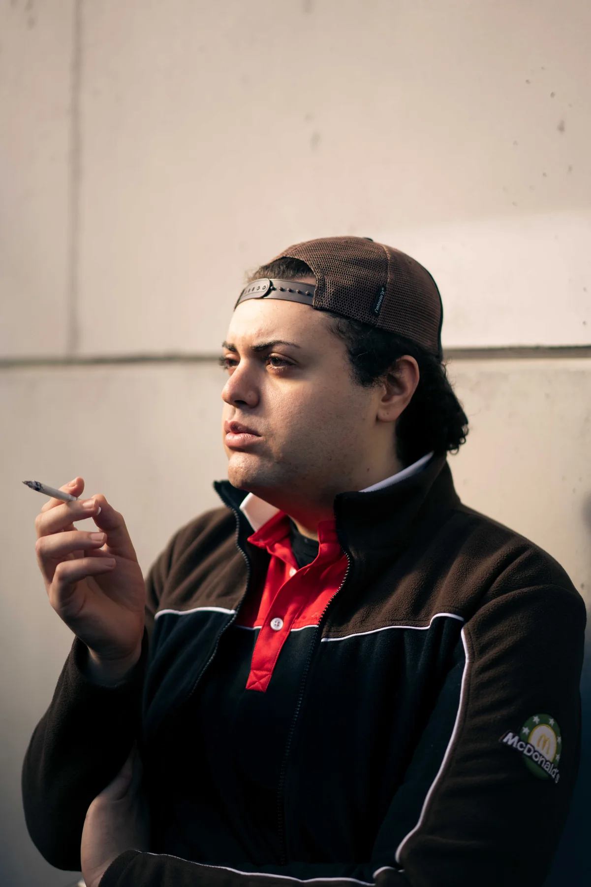 A person in a backwards cap smokes against a concrete wall.