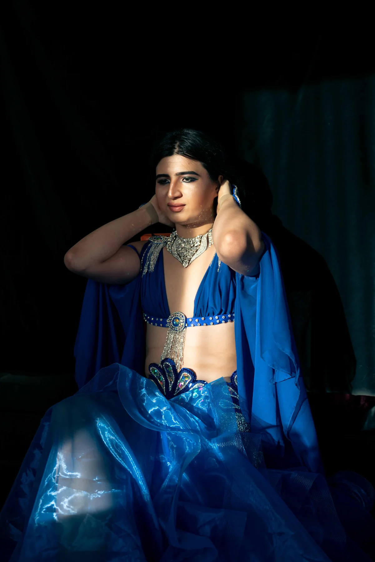 A person in a blue belly-dance costume sits in half-light, hands lifted to their hair.