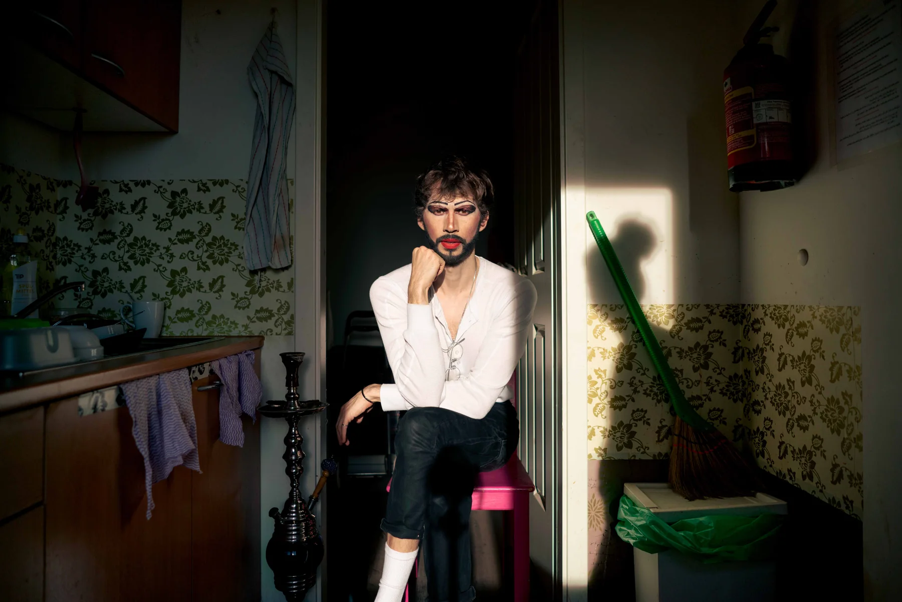 A person sits in a kitchen doorway beside a shisha pipe, white shirt open, a broom leaning in the light.