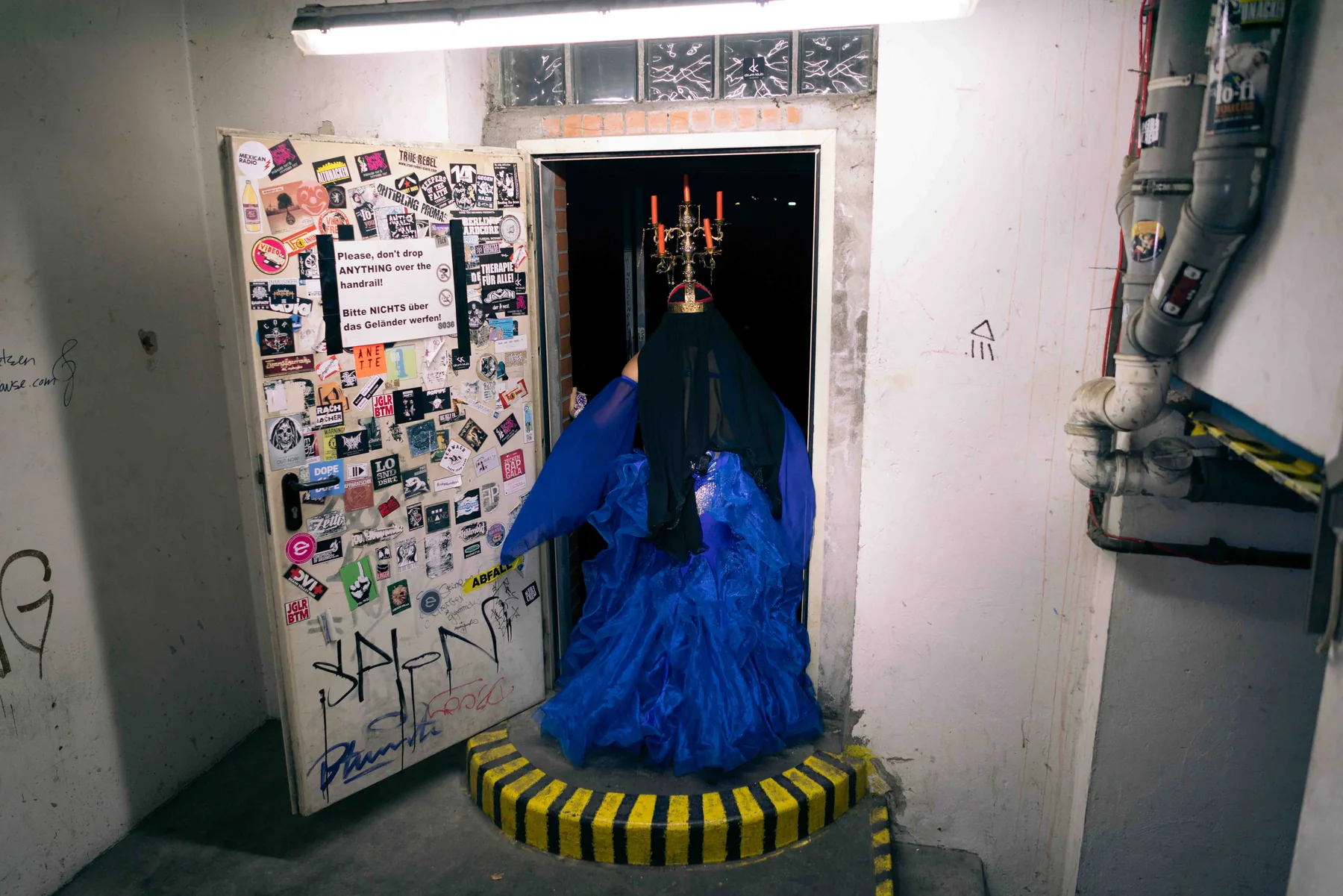 A figure in a blue gown stands in a basement doorway crowned by a candelabra.