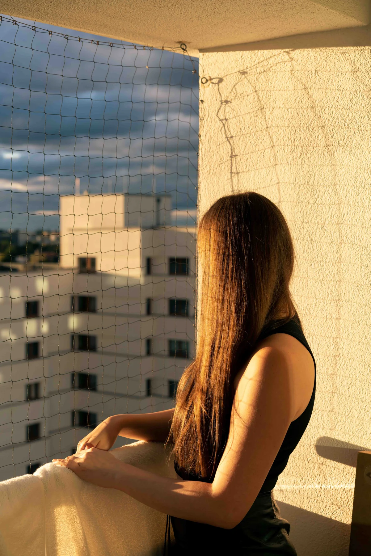 A person sits on a balcony in golden evening light, the city soft beyond the netting.