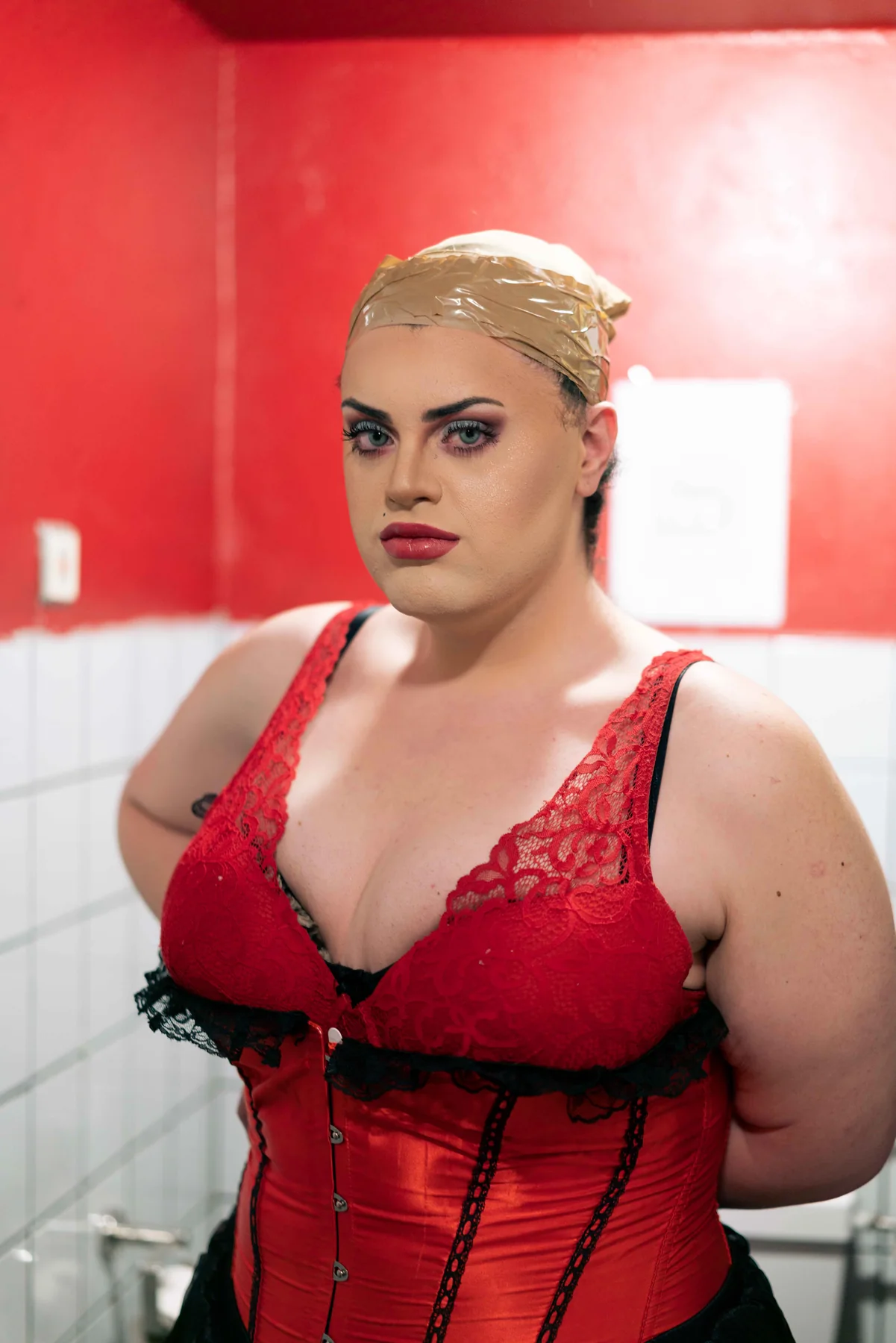 A portrait of a person in a red corset and taped wig cap against a red wall.