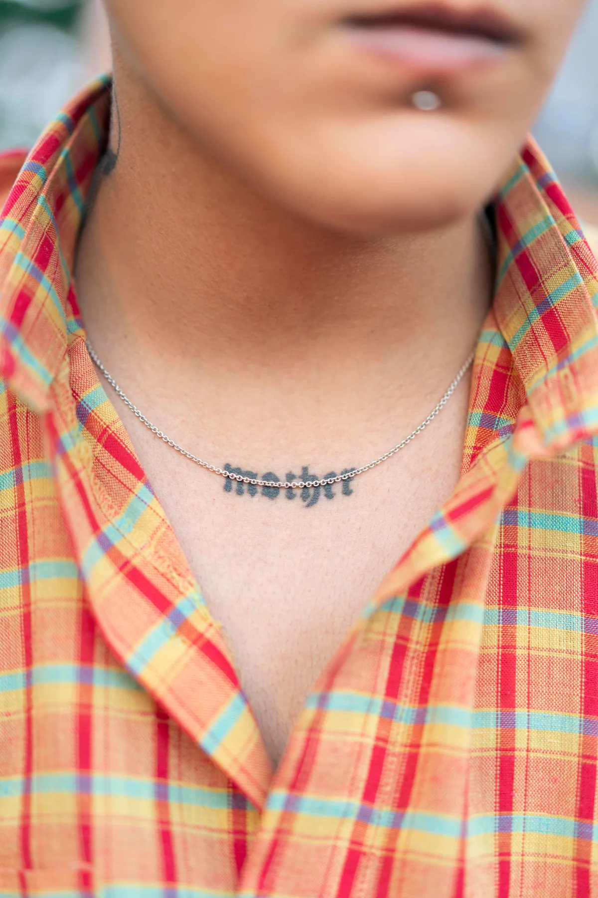 A tattoo reading “mother” at the base of a neck, under a silver chain and an open plaid collar.